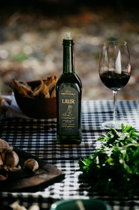 Bottle of Laur Extra Virgin Olive Oil on a checkered table cloth with a glass of red wine, empanadas and garnish